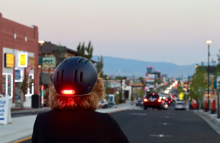 Si vede dal dietro una persona che indossa un casco nero con lucina rossa rivolta verso una strada di città con macchine